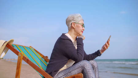 Asian elderly woman chatting with friends on video call while relaxing at the beachの写真素材
