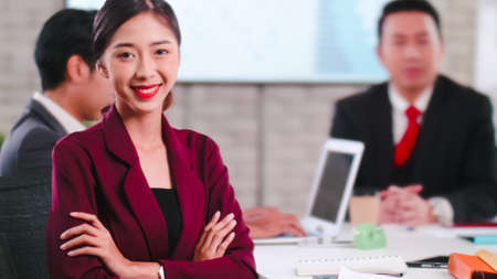 Confident young businesswoman sitting smiling at the camera in a meeting room with colleagues in the background.の写真素材