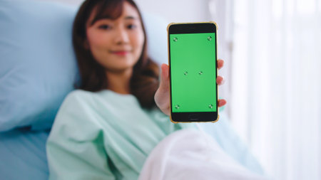 Happy young Asian female patient showing smartphone with green screen during hospitalization.の写真素材