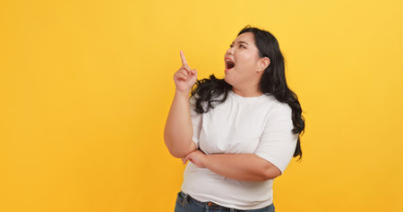 A chubby Asian woman wearing a white t-shirt is pointing at something on a yellow background.の写真素材