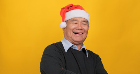 An old Asian man wearing a Santa hat smiling and looking at the camera. Isolated on yellow background in the studio.の写真素材