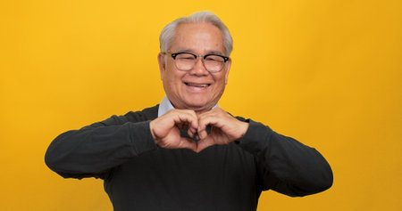 An old Asian man making a heart shape with his hands. Isolated on yellow background in the studio.の写真素材