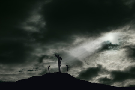 A depiction of the crucifixion of Jesus Christ on a cross with 2 other robbers nearby on Calvary. The sky is darkened with rays of light breaking through the clouds onto the cross for drama. Concept of the death of Jesus on Good Friday and His resurrectio