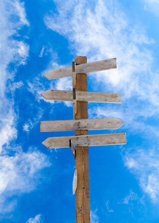 Old wooden arrow signpost against a blue cloudy sky with copy space. Idea of crossroads and concept of being lost, confused or indecisiveness. Vertical orientation.の写真素材