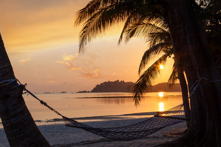 Beautiful golden sunrise over sandy beach on Bintan Island in Indonesia with silhouette of hammock and coconut palm trees with copy space.の写真素材