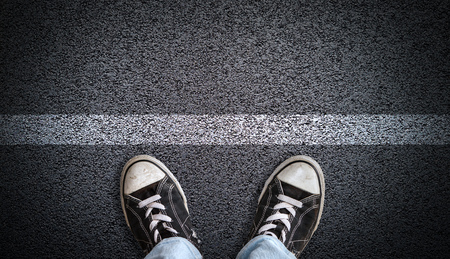 A teenager in jeans and canvas shoes standing on asphalt road behind a line with copy space. Concept of standing at a starting line, a new beginning, ready for action.の写真素材