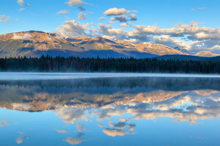 Morning sun and low clouds cover the Rocky Mountains at Annette Lake in Jasper National Park with reflections of Majestic Mountain and Aquila Mountain on the calm waters.の写真素材