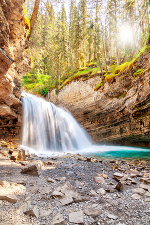 Morning sun peeks through the forest as a waterfall flows into Johnston Canyon creek at Banff National Park in Alberta, Canada. Johnston Canyon is a popular hiking place in the Canadian Rockies.の写真素材
