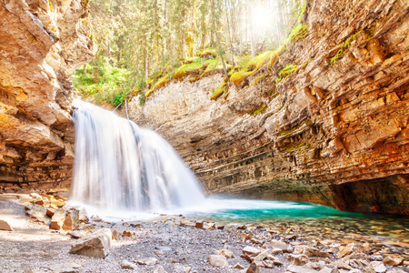 Morning sun peeks through the forest as a waterfall flows into Johnston Canyon creek at Banff National Park in Alberta, Canada. Johnston Canyon is a popular hiking place in the Canadian Rockies.の写真素材