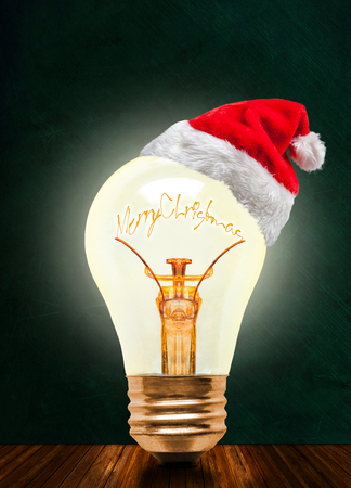 Glowing light bulb wearing Santa hat with Merry Christmas message on chalkboard background with copy space. Glowing Christmas and a bright New Year ahead.の写真素材