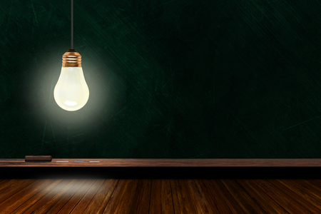 Hanging and illuminated light bulb in a classroom with background chalk board and wooden table and copy space. Concept of ideas, creativity and innovation in a learning environment.の写真素材