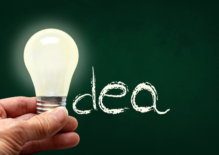 Man holding illuminated bulb with bare hands against chalkboard with the word Idea. Concept of bright idea, innovation, imagination, inspiration, vision. Copy space.の写真素材
