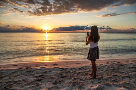 Young girl capturing vacation memories of a beautiful golden sunrise by the beach at Riviera Maya in Cancun, Mexico, with her smartphone.の写真素材