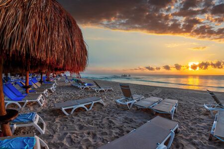 Golden sunrise over rows of lounge chairs and palm tree parasols on a sandy Caribbean beach vacation at Riviera Maya in Cancun, Mexico.の写真素材