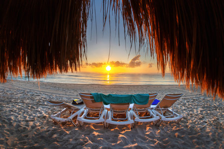 Beautiful golden sunrise over an empty beach and row of lounge chairs on the Caribbean coast of...