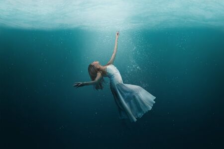 Elegant girl dancer in white dress in a state of levitation under the deep waters of the ocean with sunlight beaming on her face. の写真素材