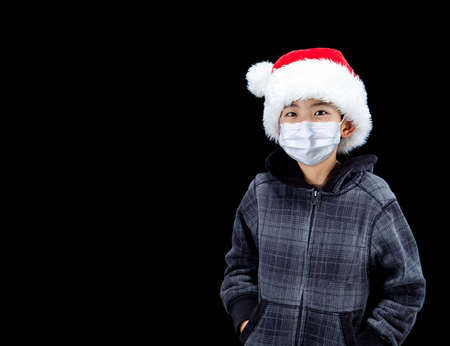 Cute boy during Covid Christmas wearing Santa hat and face mask due to pandamic, isolated on black background with copy space.の写真素材