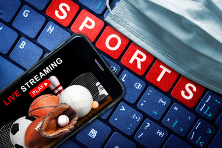 While sports venues are closed to fan attendance due to   pandemic, event Live streaming is now the new normal. Smartphone with live broadcast and medical face mask on high tech background.の写真素材