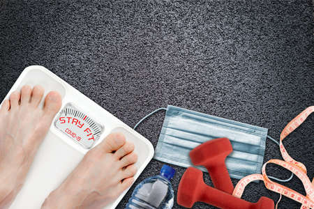 Woman on bathroom scale with Stay Fit message during  quarantine. Concept of healthy lifestyle and sport exercise during self isolation surrounded by fitness accessories, dumbbells, water bottle, measuring tape and face mask.の写真素材