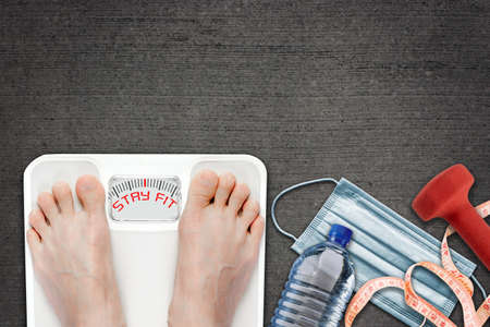 Woman on bathroom scale with Stay Fit message during  quarantine. Concept of healthy lifestyle and sport exercise during self isolation surrounded by dumbbell, water bottle, measuring tape and face mask.の写真素材