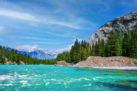 Rapidly flowing waters of Bow River in Banff National Park with surrounding pine trees and the snow-capped Canadian Rockies in the background.の写真素材