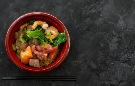 Hawaiian seafood poke bowl with diced salmon, tuna, shrimp; and salad isolated on black concrete table background with chopsticks and copy space.の写真素材