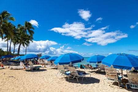 Tourists relaxing on chaise lounges with blue umbrellas on sunny Waikiki Beach in Honolulu, Hawaii.の写真素材