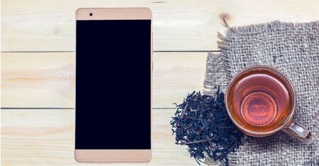 Smart phone with a cup of tea and tea leaves on wooden background.