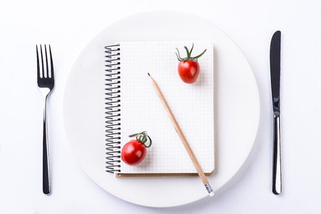 Plate, fork, knife, tomatoes, pencil and notebook tape on light wooden tableの写真素材