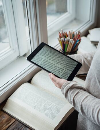 Young beautiful woman reading tablet pc and books in front of big windowの写真素材