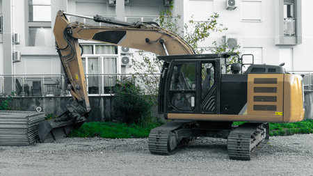 A large yellow excavator sits on a gravel lot beside a residential building, ready for earthmoving work. The hydraulic arm and bucket show active construction and development in the area.の写真素材