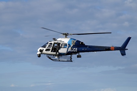 blue and white police helicopter flying in the sky, door is open and a police man is looking down