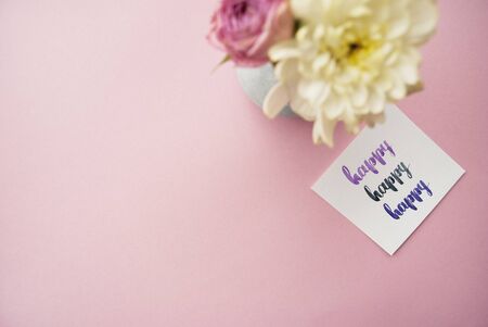"Happy" written in calligraphy style on paper with bouquet of pink roses and white chrysanthemums on a pink background. Flat lay, top viewの写真素材
