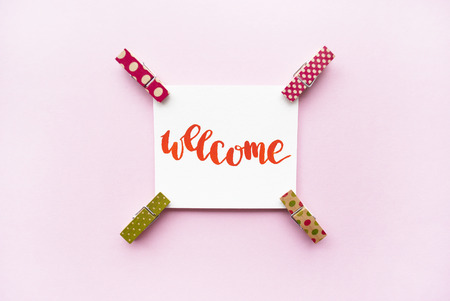 Word "Welcome" handwritten with watercolor in calligraphy style, miniature clothespins on a pink background. Flat layの写真素材
