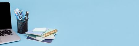Laptop, stack of notepads, stand with stationery on a blue background. Banner with workspace concept.の写真素材