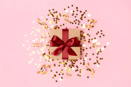 Gift box tied with red ribbon on a pink background with gold stars confetti. Festive composition.