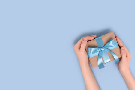 Female hands hold gift box on a blue background. Present tied with a ribbon. Festive composition with place for text.の写真素材