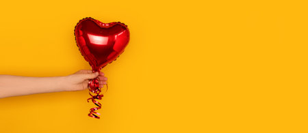 Banner with hand hold red inflatable foil balloon in a heart shape. Festive concept in front of yellow background with copy space. selective focus.の写真素材