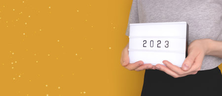 Banner with female hands hold lightbox with 2023 numbers in front of yellow background. New Year...