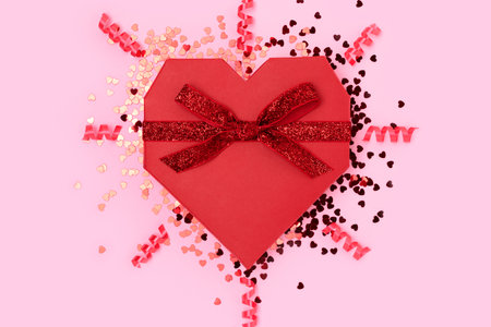 Red gift box in a heart shape tied with glittering ribbon. Festive concept with scattered confetti on a pink background.の写真素材