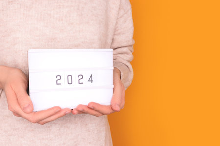 Female hands hold lightbox with 2024 numbers in front of yellow background. New Year concept with place for your design.の写真素材