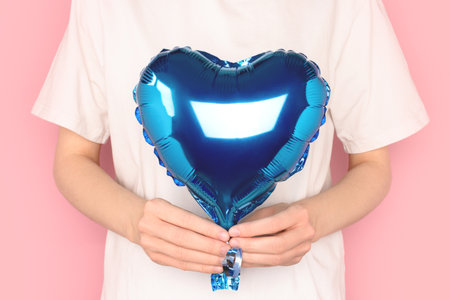 Woman in a white t-shirt hold in hands blue foil balloon in a heart shape. Holidays concept in front of pink background.の写真素材