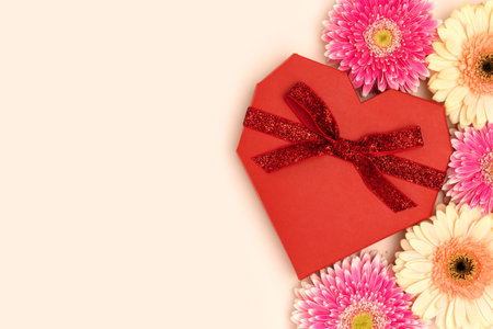 Red gift box in a heart shape with a tied bow and gerbera flowers on a beige background.の写真素材
