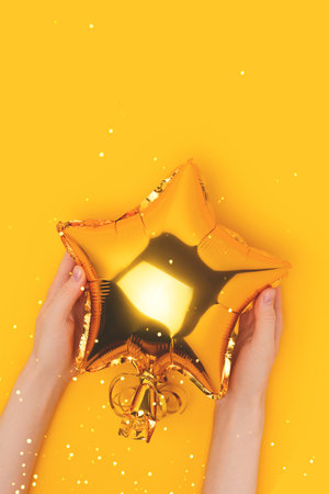 Female hands hold golden foil balloon in a star shape. Creative composition on a yellow background.の写真素材