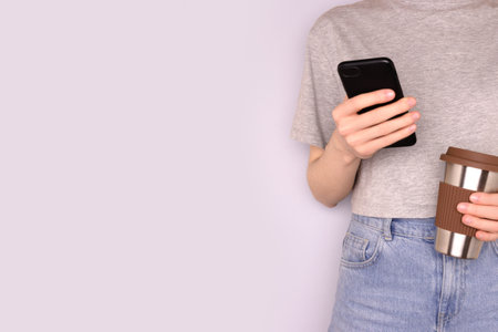 Woman in a gray t-shirt hold in hands coffee cup and use smartphone in front of blue background. Selective focus.の写真素材