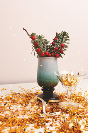 Nobilis fir and winterberry branches in a vintage vase, small crystal glass and gold confetti in front of beige background. Festive composition.の写真素材