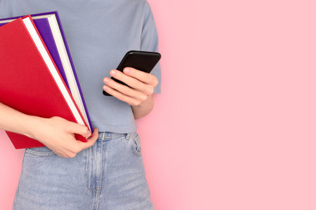 Woman holding books in her hands and using smartphone in front of pink background. Place for text.