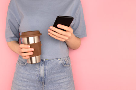 Student girl in a blue t-shirt hold in hands coffee cup and use smartphone in front of pink background. Selective focus.の写真素材