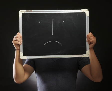 unhappy face drawn on a blackboard holding by a young man in photography studio with black backgroundの写真素材