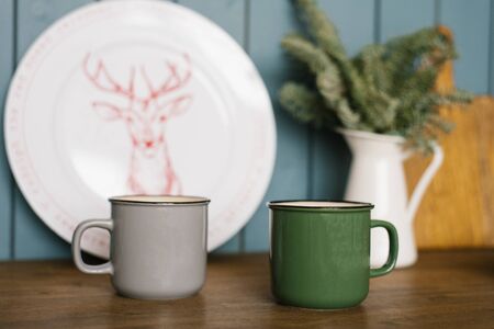 metal mugs of gray and green are on the countertop of the kitchen, decorated for Christmas and new year. In the background a white plate with a deer and a bouquet of spruce in a white vaseの写真素材